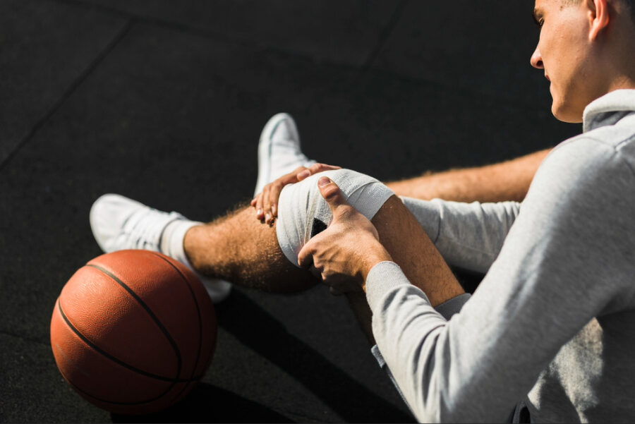 Athlete sitting on outdoor court holding an injured knee wrapped in a bandage next to a basketball, illustrating sports injury, pain, and rehabilitation concerns.