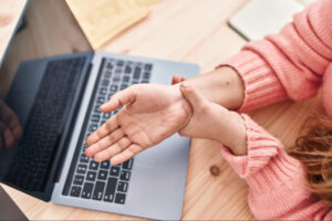 Person holding their wrist in pain while using a laptop, illustrating common symptoms of carpal tunnel syndrome such as wrist discomfort, numbness, and repetitive strain.