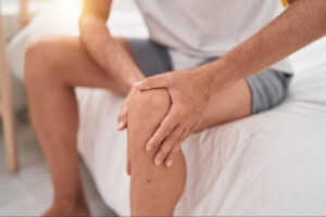 Man holding his knee in discomfort while sitting on a bed, representing pain, swelling, or warning signs after knee arthroplasty surgery recovery.